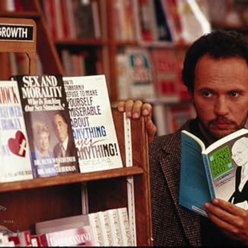 Billy Crystal holding a book and standing behind a book shelf looking off into the distance.
