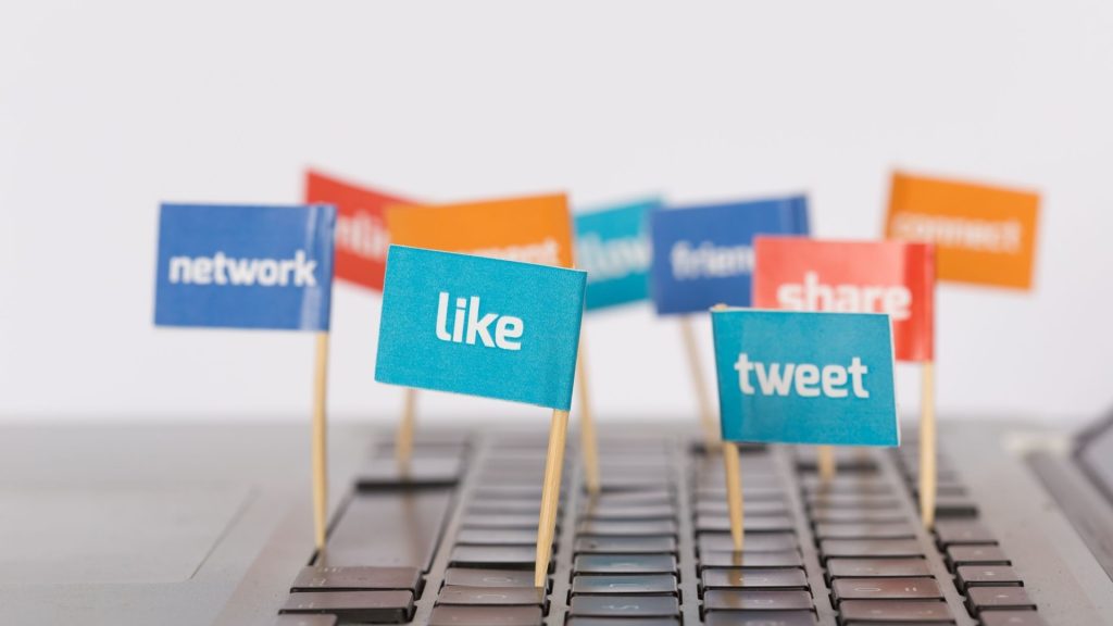 A laptop keyboard with flags sticking out from between the keys of varying colors with words like "network", "like", "tweet", and "share" on the flags.