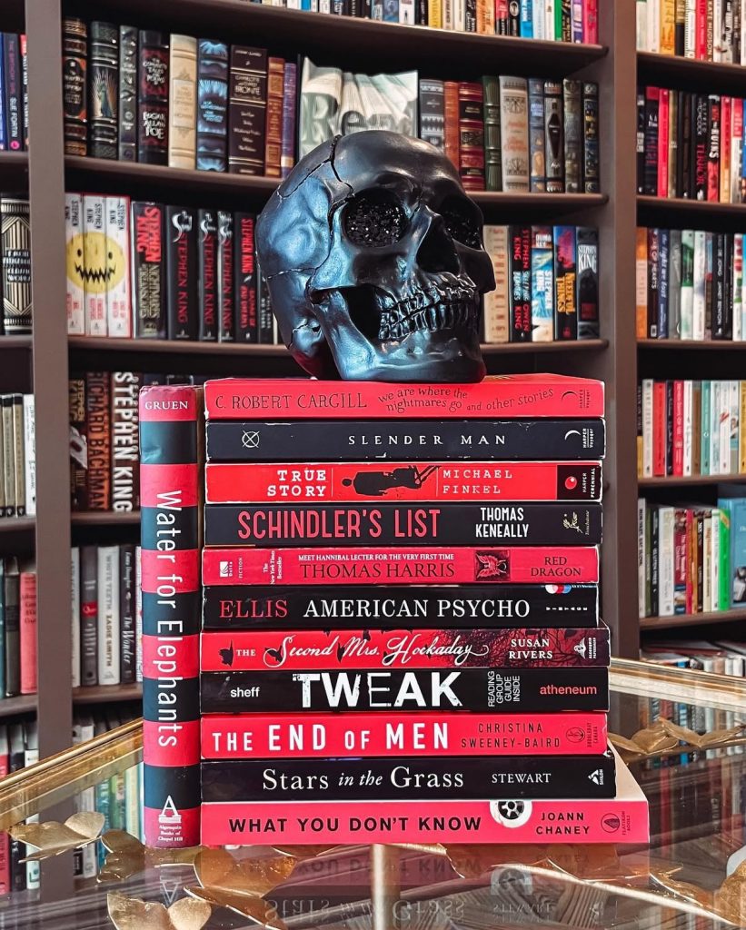 Neatly stacked black and red books placed on a desk in an alternating patter with the spines facing toward the camera, including the titles Slender Man, Schindler's List. An all black skull with black bedazzled eyes is placed on top of the stack as decoration.