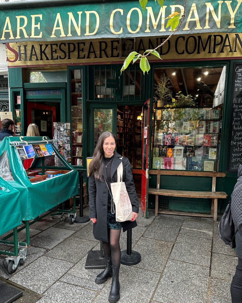 Bookstagrammer Laura, @storiesandskulls, standing outside of the Shakespeare and Company Bookstore. She wears a long black trench coat with a black undershirt, a navy blue floral skirt, black tights, and leather boots. She carries a tote bag from the bookstore.