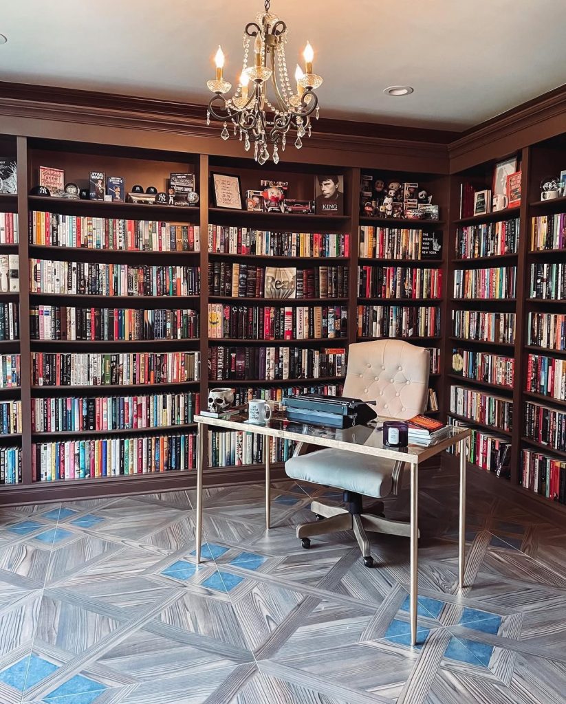 The library Laura created in her home containing book shelves that span from ceiling to floor and are filled with books of all genres. There is a desk in the corner with a swivel chair; on the desk is a skull, typewriter, mug, candle, and a stack of books.
