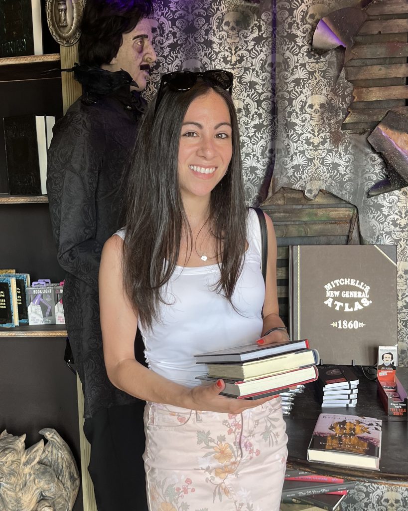 Bookstagrammer Laura, @storieandskulls, holding three books in front of a Edgar Allen Poe statue located in a horror bookstore. She is wearing a white tank top and floral jeans with blank sunglasses on her head.