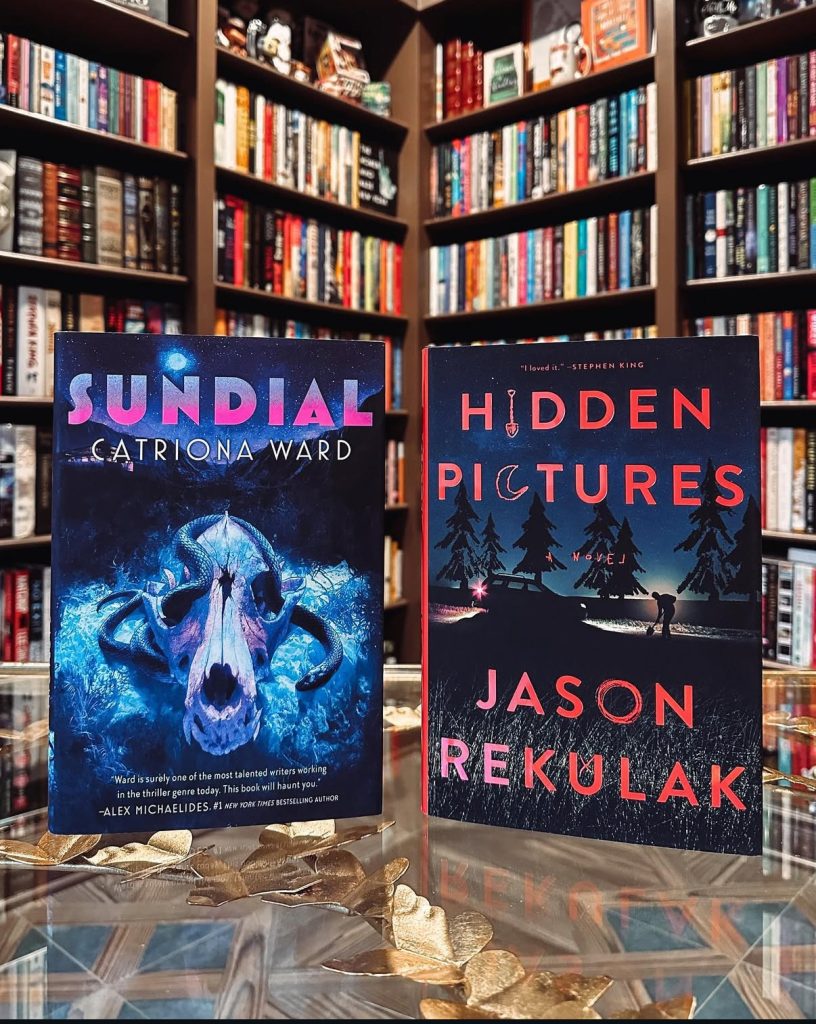 Book cover for Sundial by Catriona Ward depicting a snake slithering through an horse's skull under the blue moonlight. Book cover for Hidden Pictures by Jason Rekulak depicting a silhouette of an SUV on the side of a road with a man in front of it digging and trees in the background. The background of this photo is book shelves filled with books.