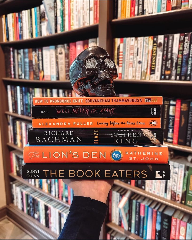 Bookstagrammer Laura holds a stack of orange and black books in an alternating pattern. The stack includes The Book Eaters and The Lion's Den among others. On top of the stack is a shiny black skull with silver bedazzled eyes. The background of the image is Laura's bookshelves.