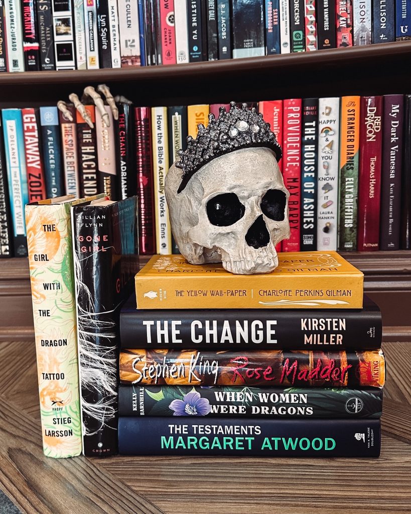 Stack of books including The Yellow Wall-paper, Gone Girl, and The Girl With The Dragon Tattoo, with a decorative skull wearing a tiara on top of them. In the background there are neatly shelved books with a decorative skeleton hand creeping over three of the book spines.