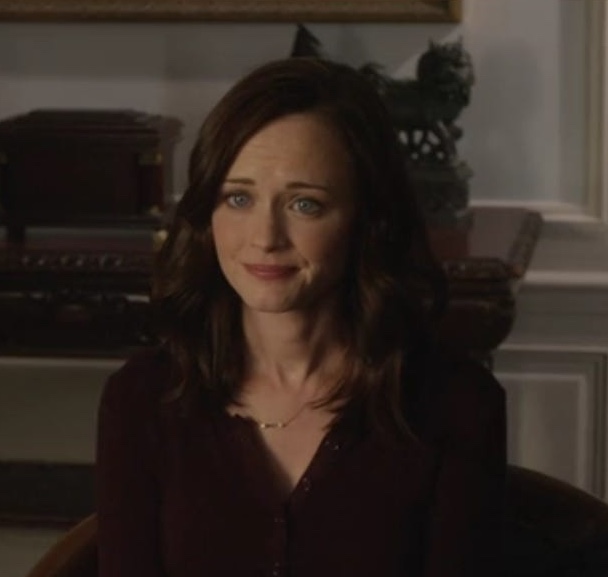 A still from the show Gilmore Girls: A Year in the Life. A woman with pale skin, blue eyes, and wavy brown hair wears a maroon button-up sweater and sits in a brown chair. Behind her is a wooden buffet table with fancy decor.