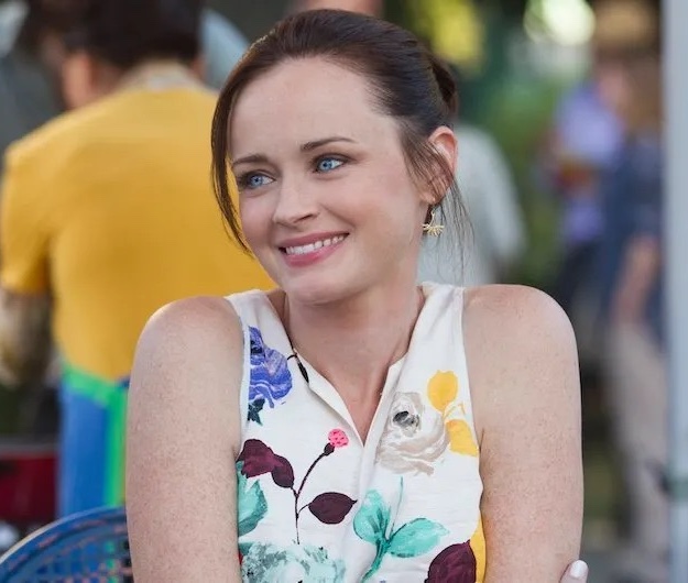 A still from the show Gilmore Girls: A Year in the Life. A woman with pale skin, blue eyes, and brown hair pulled into a bun smiles at something off camera. She wears a sleeveless white blouse with multicolored flowers.