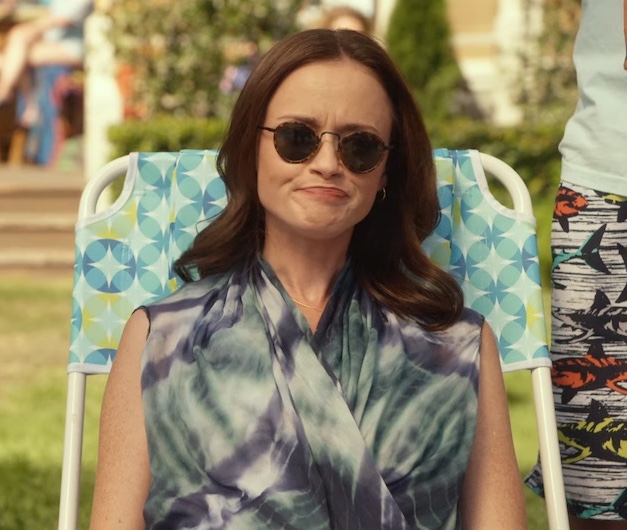 A still from the show Gilmore Girls: A Year in the Life. A woman with pale skin and wavy brown hair wears round sunglasses and has a frustrated expression. She sits on a blue printed pool chair and wears a sleeveless top with a teal and purple tie dye pattern.