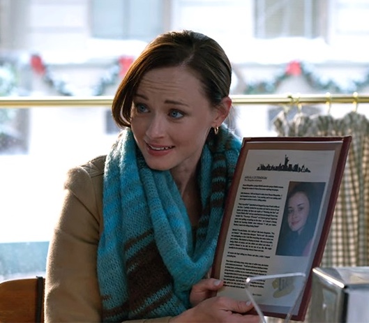 A still from the show Gilmore Girls: A Year in the Life. A woman with pale skin, blue eyes, and pulled back brown hair wears a tan coat and a blue stripes scarf. She sits at a table in a diner and incredulously holds up a menu that features a printed article beside a picture of her younger self.