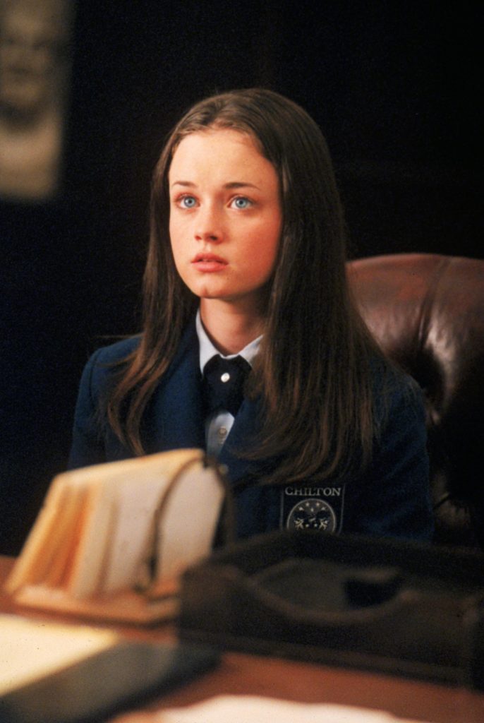 A still from the show Gilmore Girls. A young woman with long brown hair, blue eyes, and pale skin, sits in a leather armchair and wears a blue private school uniform.