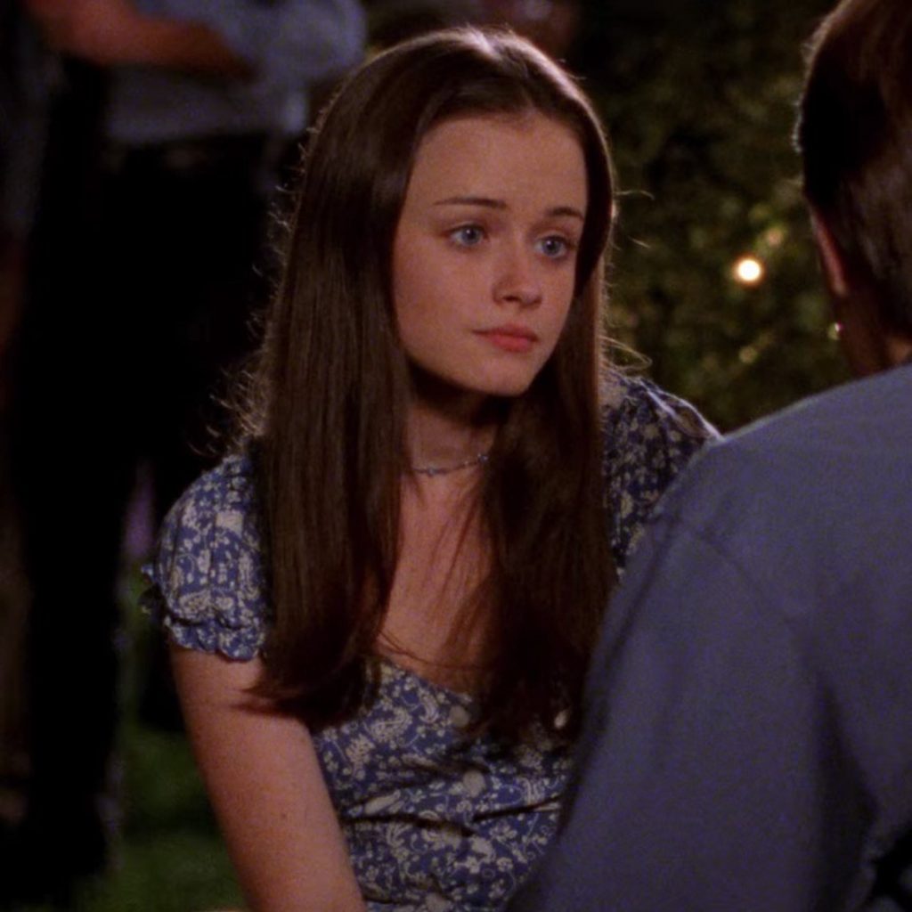 A still from the show Gilmore Girls. A young woman with pale skin, long brown hair, and blue eyes wears a blue floral dress and looks at a young man seated beside her.