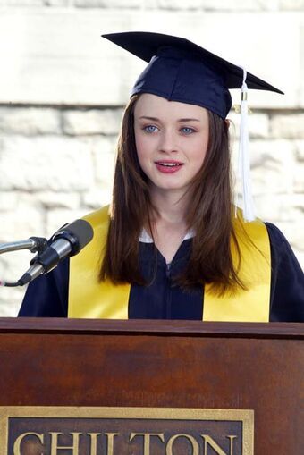 A still from the show Gilmore Girls. A young woman with pale skin, long brown hair, and blue eyes stands behind a wooden podium and wears a blue graduation gown with a white tassel and a gold Valedictorian stole.