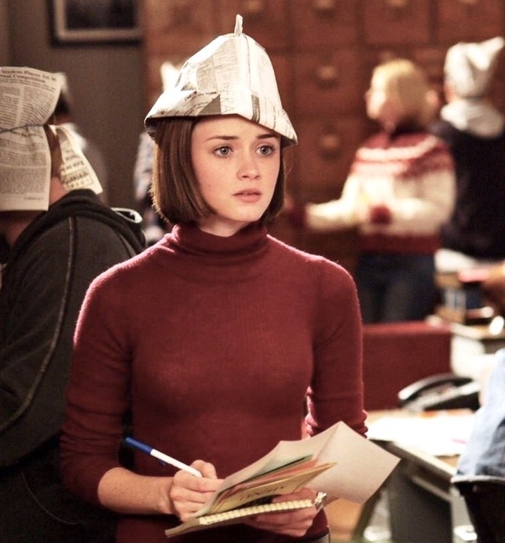 A still from the show Gilmore Girls. A young woman with short brown hair, pale skin, and blue eyes wears a red turtleneck sweater and a newspaper hat. She holds a pen and series of papers and stands in a writer's room, surrounded by other young men and women, some also wearing newspaper hats.