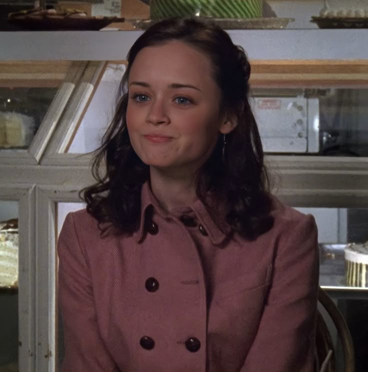 A still from the show Gilmore Girls. A young woman with curly brown hair, pale skin, and blue eyes wears a pink coat and smiles. Behind her is a glass counter display full of cakes.