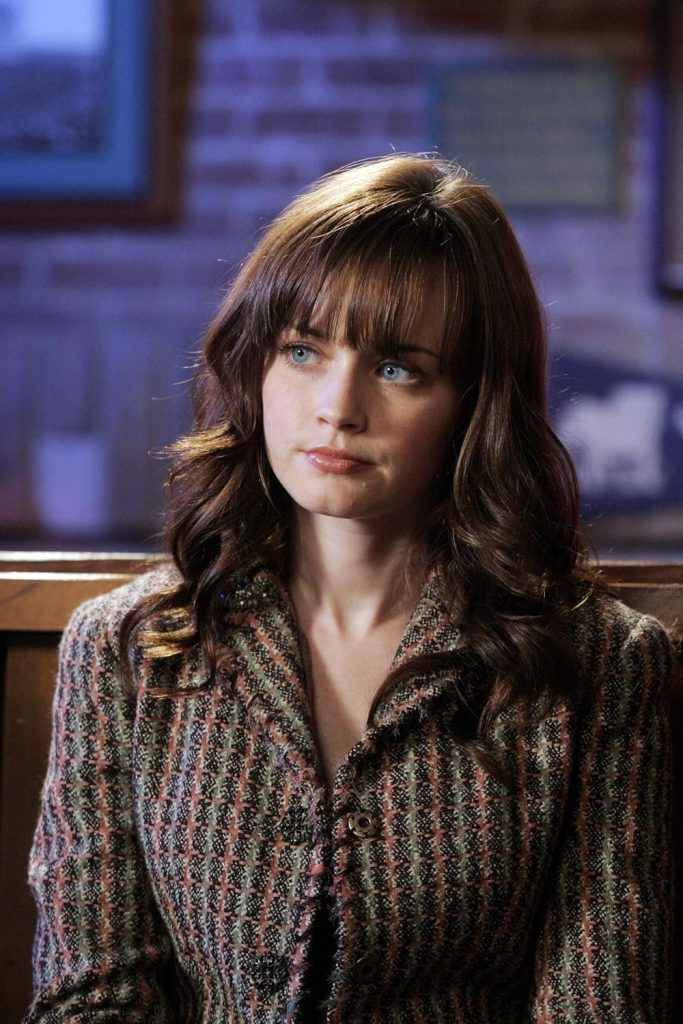 A still from the show Gilmore Girls. A young woman with pale skin, blue eyes, and long curly brown hair with straight bangs wears a brown, green, and pink tweed blazer and sits in a wooden restaurant booth. The back wall is lit with a blue light.