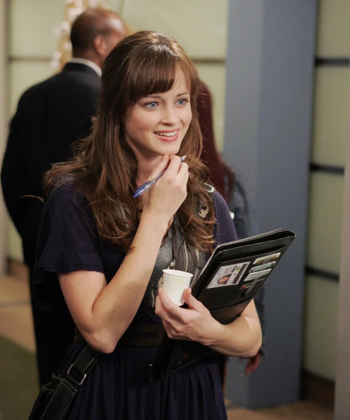 A still from the show Gilmore Girls. A young woman with pale skin, blue eyes, and curly brown hair with straight bangs stands in a hallway and smiles. She holds a pen, a black padfolio with an ID badge, and a small white plastic cup.
