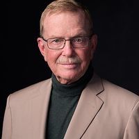 Dr. Randolph B. Schiffer wears a gray turtleneck shirt underneath a tan blazer and a pair of rectangular black wire glasses. He has short blonde hair and a short white mustache.