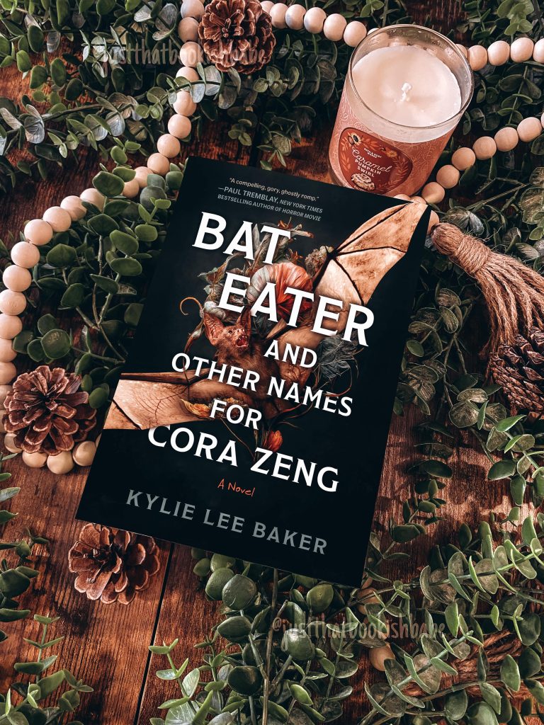 Bat Eater And Other Names For Cora Zeng by Kylie Lee Baker book cover on a wood table surrounded by decorative green leaves, scattered pine cones, one lit candle, and a strand of lightly-colored wooden beads.