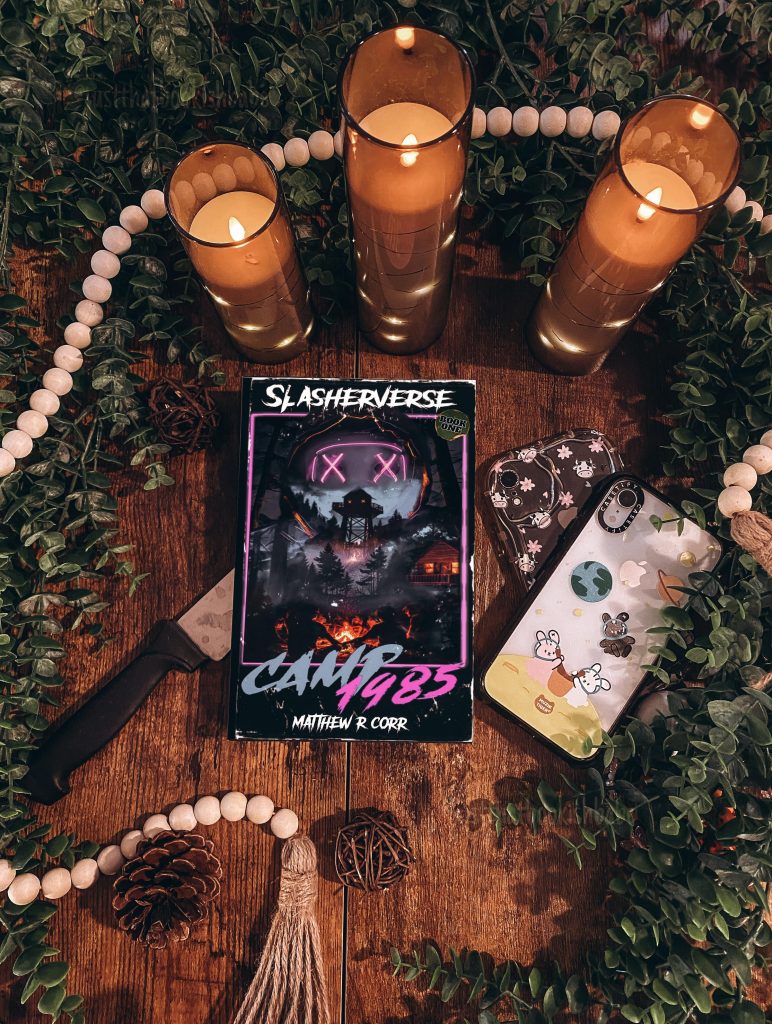 Camp 1984 (Slasherverse) by Matthew R. Corr book cover laid flat on a knife on a wooden table with a phone nearby. Above the book cover are three lit candles and it is surrounded by decorative green leaves and a strand of large white beads.