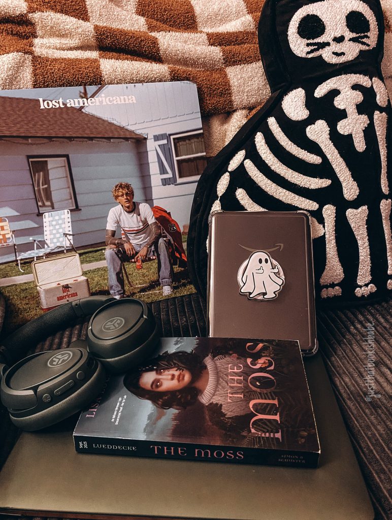 THe Moss by Lisa Lueddecke book cover lying on a glassy table with a covered kindle, headphones, a skeleton throw pillow, and the vinyl record cover for Lost Americana by MGK nearby.