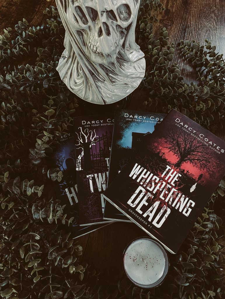 The Gravekeeper books by Darcy Coates fanned out on a wooden table surrounded by decorative greenery with a shrouded skull on the table above them.