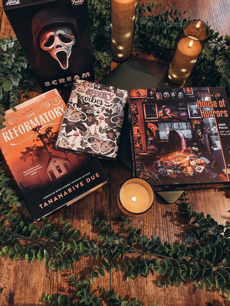 The Reformatory by Tananarive Due and The House of Horrors by Suzanne Weyn book covers laid on a wooden table surrounded by decorative green leaves, three lit candles, scream mask decor, and a kindle cover with moth stickers on it..