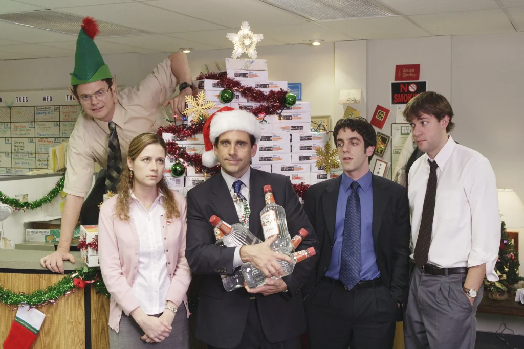 The Office, Christmas Party. At reception episode still. Michael (Steve Carell) wears a Santa hat and holds five bottles of vodka, Jim (John Krasinski) stares at him uncomfortably, Ryan (B.J. Novak) stands next to him irritated, Pam (Jenna Fisher) rolls her eyes, and Dwight (Rainn Wilson), in an elf hat, climbs on the desk behind them. They stand in front of a tree made of paper packages.