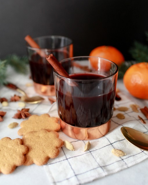 Swedish glogg mulled wine served in glass cups with cinnamon sticks, accompanied by traditional gingerbread cookies, fresh oranges, and scattered spices including star anise and almonds on a white kitchen towel.