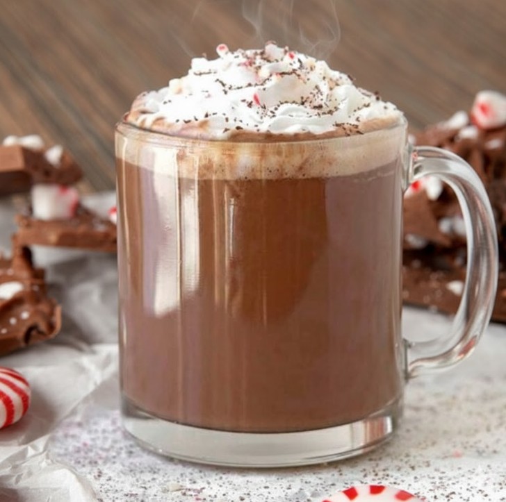 Peppermint white chocolate mocha in a clear glass mug topped with whipped cream, crushed candy canes, and chocolate drizzle. The festive holiday drink is surrounded by homemade peppermint bark and scattered peppermint candies.