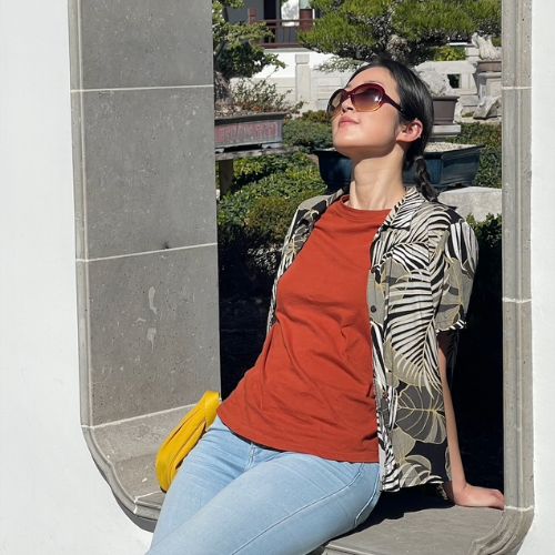 Author, Christine Han, wearing brown sunglasses, a monochrome floral patterned shirt with a red top underneath, and blue jeans, sitting with her hands behind her back, leaning backwards and looking above.