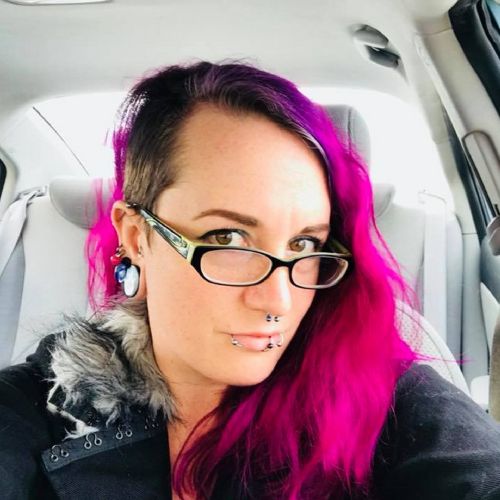 Author, Augusta Wickman, wearing a black jacket with white fur hoodie, black-yellow rectangular glasses, red-blue-white earrings, with nose and lip piercings, sitting in a car with white seats, looking ahead.
