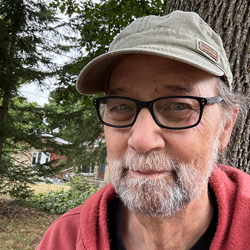 Photograph of author Charles de Lint. De Lint looks into the camera smiling, with several trees and a house behind him.
