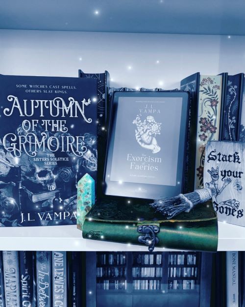 A white bookshelf stacked with books, including a physical copy of Autumn of the Grimoire by J.L. Vampa next to a crystal, a tablet displaying J.L. Vampa's The Exorcism of Faeries stacked atop another leather-bound book, and a wooden sign reading "Stack your Bones" with a skeleton wearing a crown. The image is tinted in grayish blue.
