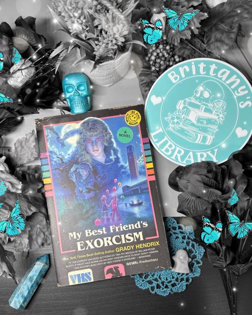 Book flatlay featuring My Best Friend's Exorcism by Grady Hendrix laid out along side flowers, teal butterflies, a teal skull, a teal-tinted crystal, a ghost, a lace doily, and a teal and white circular pin reading "Brittany's Library" featuring a skull and flowers laid atop a stack of books. Except for the book cover and teal accents, the image is black and white.