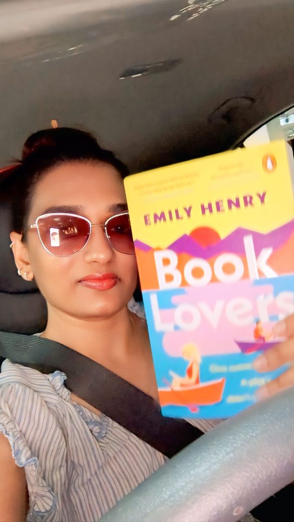 Bookstagrammer Yesha smiles for a selfie in the car while holding up Emily Henry's Book Lovers. Yesha wears sunglasses, a neat bun, and a striped shirt.