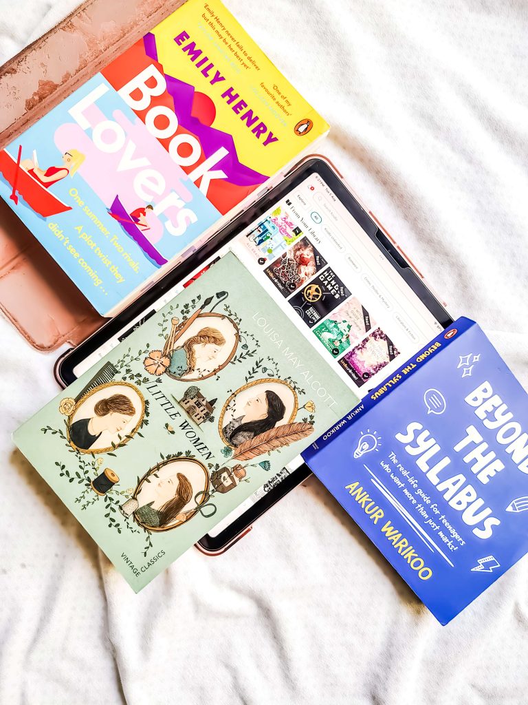 On top of an iPad, open to Yesha's Goodreads page, is a sage edition of Louisa May Alcott's Little Women. To the right is Ankur Warikoo's Beyond the Syllabus and to the left is Emily Henry's Book Lovers. These objects rest on a bed with white sheets.