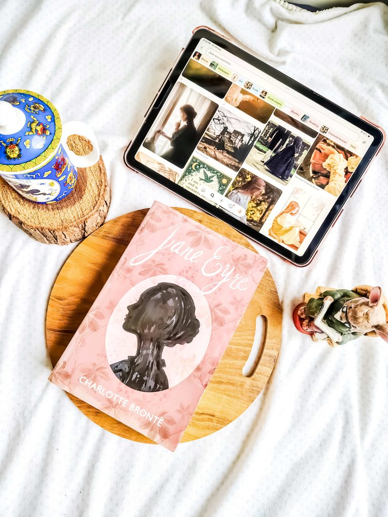 On top of a wooden board is a pink edition of Jane Eyre by Charlotte Bronte. A coaster made of a tree branch with a colorful teacup sits next to it as well as a little woodland figurine reading a novel. In front of Jane Eyre is Yesha's iPad displaying images aesthetically similar to Bronte's novel.
