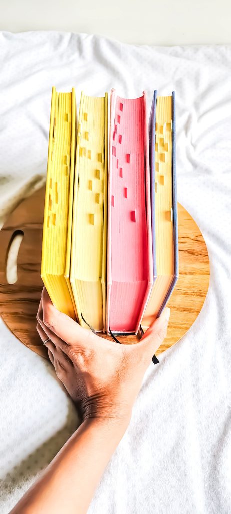 Yesha's hand holds four hardcover novels, with their page sides up on top of a circular wooden board. The first two novels have yellow bindings and pages, the third has a pink binding and pink pages, and the last novel has a purple binding with yellow pages.