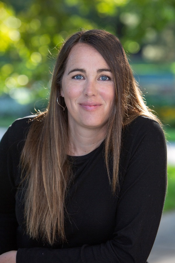 Bianca Schulze author headshot shows Bianca standing in front of a leafy, sunny blurred out background. She props her left arm on something that's out of sight. She wear a long sleeve, black crewneck shirt and her long, brunette hair is parted to the side and goes around one of her shoulders. Her blue eyes can be seen with silver hoop earrings. She gives a closed-lip smile at the camera.