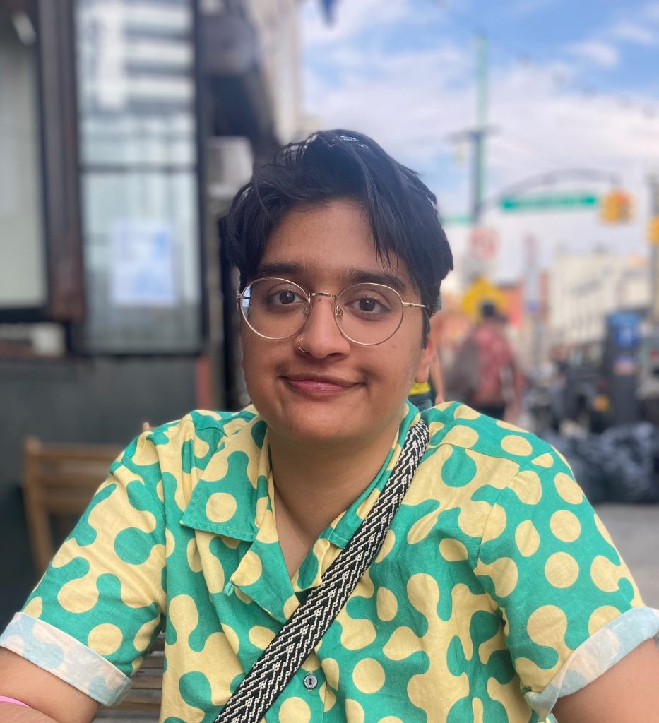 Jehan Madhani author headshot shows Jehan sitting in a chair on some sort of outside patio in a bustling city. She wear a bright green and muted yellow button up shirt with the short sleeves cuffed at the ends. Her hair is in a short, black haircut with a side part. She wears gold-rimmed frame glasses as she gives a closed-lip smile at the camera.
