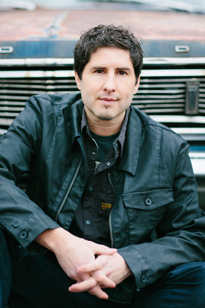 Matt de la Pena author headshot shows Matt sitting in front of the grill of a classic-looking car. He wears a dark, black jacket with a similar dark button-up underneath. Matt has short, black hair with brown eyes. He holds his hands clasped in front of him as he looks into the camera with a serious look.