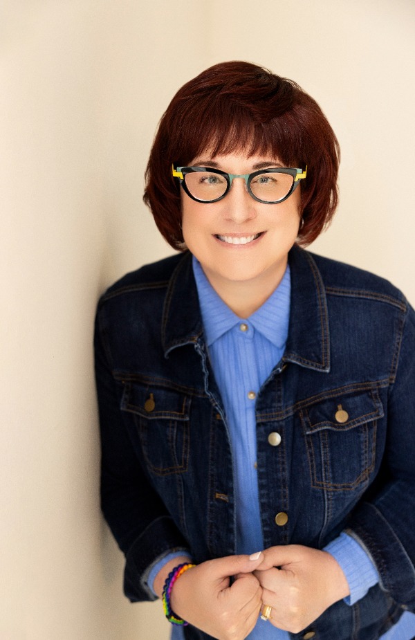 Pat Zietlow Miller author headshot features the author leaning against a tan wall with her right shoulder in a dark blue denim jacket and a light blue button-up shirt underneath. She has short, brown hair with bangs and wears black frame glasses. She smiles warmly at the camera.
