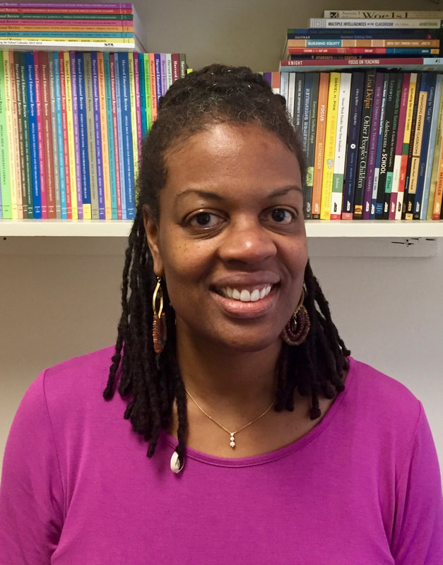 Valerie Bolling stands in front of a bookshelf with different colors and size books. She gives the camera a welcoming smile with her dark hair tied in a half-up style with a bun in the back. She has a bright, purple crewneck shirt on and a pretty gold necklace paired with long, golden earrings.