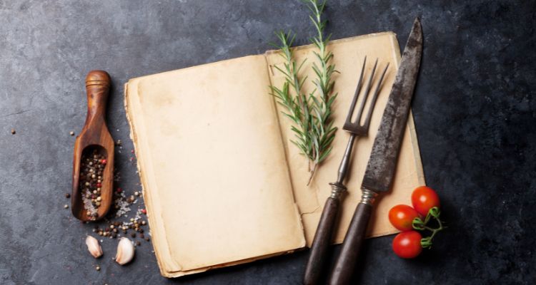 This image features an open book with a fork and knife, three tomatoes, a sprig of rosemary, and a spice spoon on a gray table.