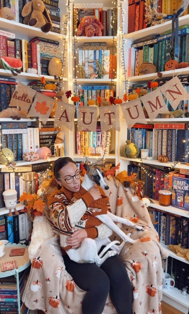 Bookstagrammer Laura Patricia Rose holding a greyhound dog, both wearing fall sweaters, sitting in a cozy reading chair. She's in her library reading nook that is decorated with fairy lights, jelly cat plushies, and a banner that reads "hello autumn."