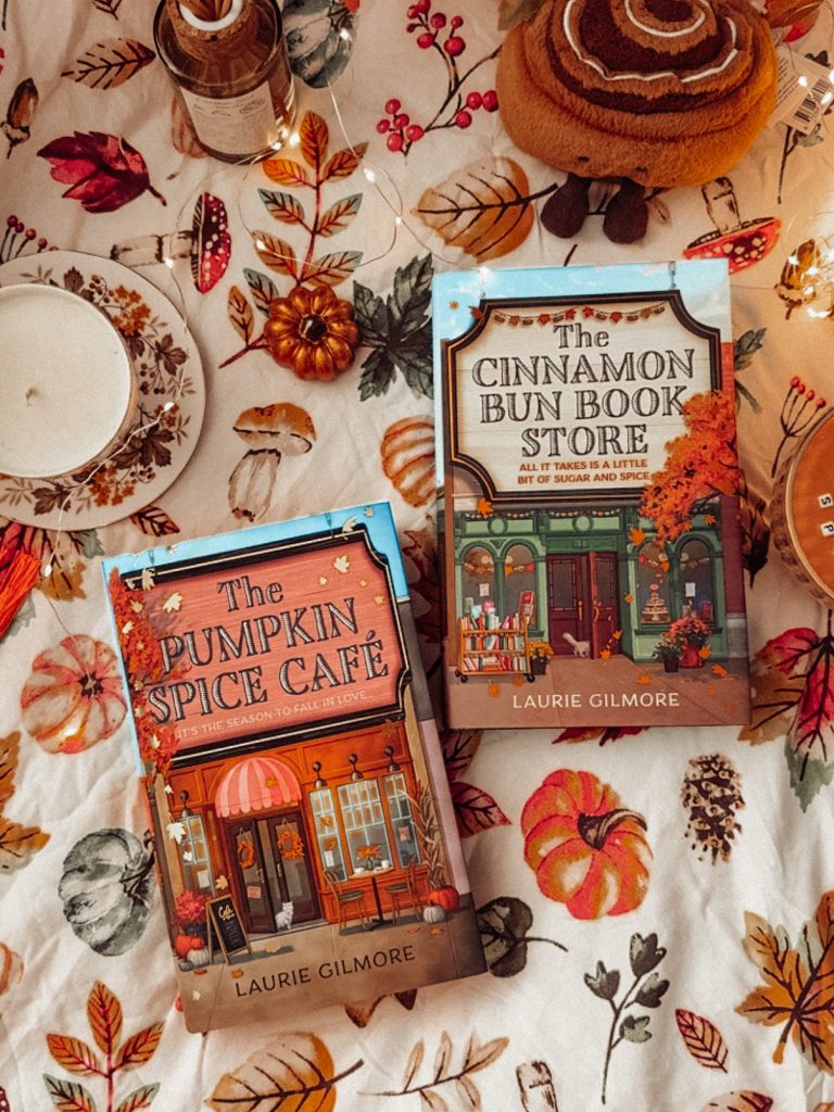 The Pumpkin Spice Cafe and The Cinnamon Bun Book Store, two cozy Fall romance novels by Laurie Gilmore, photographed by Bookstagrammer Laura. In the shot, the books are placed on a pumpkin, leaf, and mushroom blanket with a candle and jelly cat used as decoration to set the cozy mood.