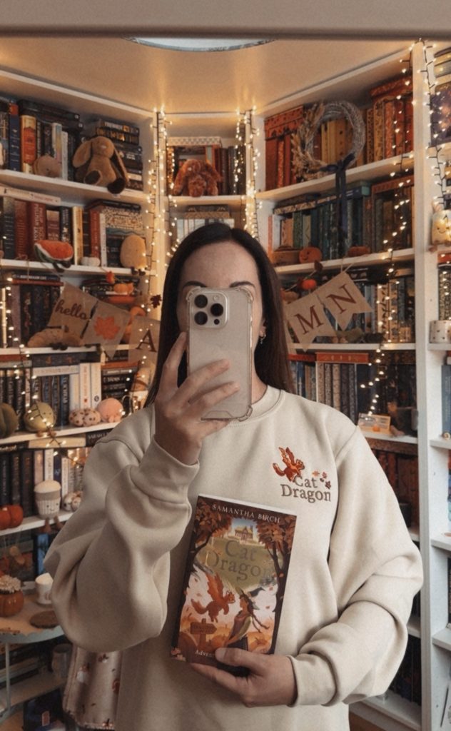 A mirror selfie captured by Bookstagrammer Laura of her holding the novel Cat Dragon by Samantha Birch, while wearing her Cat Dragon merchandise crew neck. She took the photo in her home library reading nook that is decorated with fairy lights, jelly cat plushies, and a banner that reads "hello autumn."
