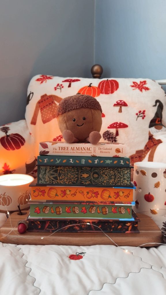 A themed book stack photographed by Bookstagrammer Laura showing off books with festive autumn sprayed edges, including visuals of leaves, pumpkins, pinecones, tree branches, and deer. An acorn jelly cat plush sits on top of the books with a lit pumpkin candle in the background, a large fuzzy fall pillow, and a ceramic pumpkin jar.