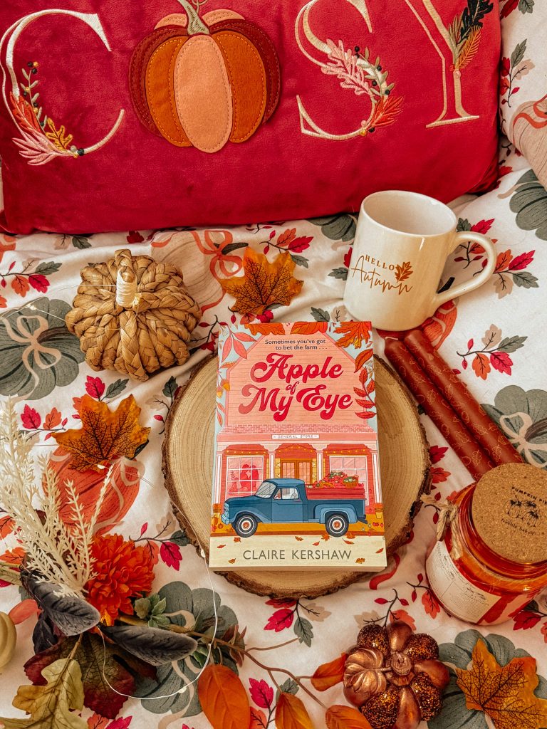 Apple of My Eye, a romance novel by Claire Kershaw, photographed by Bookstagrammer Laura with an autumn aesthetic background. Decorations include a throw pillow that says cozy, a woven pumpkin, orange candles, a tree trunk disc the book is placed on, a mug that reads, "hello autumn" all placed on top of a pumpkin throw blanket.