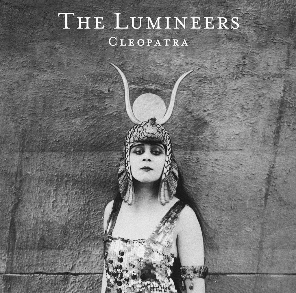 The album cover for Cleopatra by The Lumineers. In black and white, a pale-skinned woman with black hair wears an ancient horned headdress, a shiny dress, and a jeweled arm band and stands in front of a rock wall.